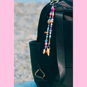 Peut inclure: Un sac à dos noir doté d'un porte-clés décoratif en perles. Le porte-clés se compose de deux brins de perles colorées dans des tons rose, bleu, violet et transparent, agrémentés de divers petits coquillages naturels suspendus à un anneau argenté.