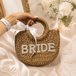 May include: A round woven straw handbag featuring the word BRIDE in large, white, pearl-encrusted block letters across the front. The bag has a circular handle and is decorated with a white satin ribbon tied in a bow on the left side.