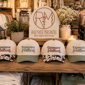 May include: Four baseball caps arranged in a row on a wooden shelf. Each cap features a cream-colored crown with the text Expensive & Difficult embroidered in brown. Two caps have cow-print brims, while the other two have solid olive green brims.