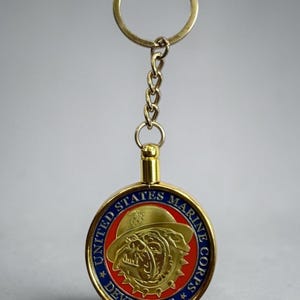 May include: A circular gold-toned metal keychain featuring a central embossed bulldog wearing a helmet against a red background. The blue outer border displays the text UNITED STATES MARINE CORPS and DEVIL DOGS with two small gold stars.