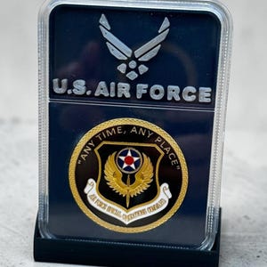 May include: A rectangular display case containing a U.S. Air Force emblem at the top and a circular Air Force Special Operations Command challenge coin below. The coin features a gold winged sword, a central star emblem, and the text ANY TIME, ANY PLACE.