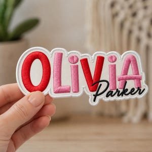 Może przedstawiać: Personalizowana naszywka z wyhaftowanym imieniem Olivia w czerwono-różowych literach blokowych z białą obwódką. Imię Parker jest wyhaftowane czarną kursywą pod literami i, v oraz i, na białym podkładzie z tkaniny.