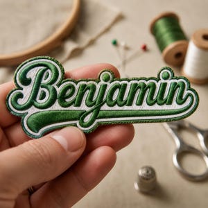 May include: An embroidered patch featuring the name Benjamin in a bold, cursive script. The lettering is dark green with a white outline and a thick green border, designed in a classic athletic style with a prominent underline flourish.