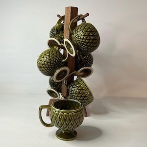 May include: A set of olive green ceramic mugs with a raised diamond grid pattern, each featuring a pedestal base and a handle. The mugs are displayed on a vertical wooden rack with multiple pegs, with one mug placed in front on the surface.