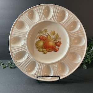 May include: A round white ceramic egg serving platter featuring twelve recessed oval indentations arranged in a circle around a central basin. The center of the platter is decorated with a colorful illustration of a pear and gooseberries.