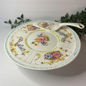 May include: A round ceramic cake stand and matching server featuring a cream-colored base with embossed scrollwork and hand-painted floral motifs in shades of pink, blue, yellow, and green. The edges are decorated with a light blue dotted pattern.