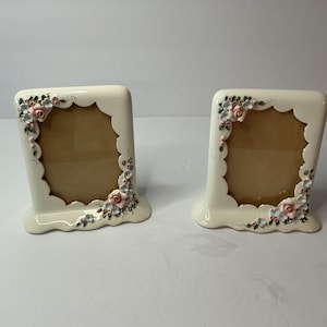 May include: A pair of off-white ceramic picture frames with a scalloped inner edge. Each frame features hand-painted floral accents, including small pink roses and tiny blue flowers with green leaves, positioned at the top left and bottom right corners.
