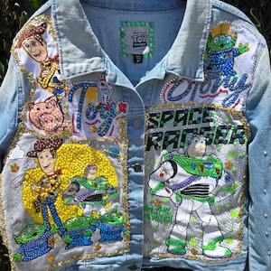 May include: A light blue denim jacket featuring Toy Story character patches and rhinestone embellishments. The jacket displays Woody, Buzz Lightyear, and the green aliens, with text reading Toy Story and Space Ranger. The collar has green beads.