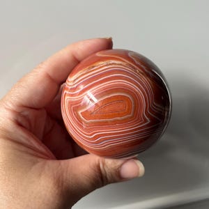 May include: A polished spherical stone featuring concentric bands of burnt orange, terracotta, and white. The surface is smooth and reflective, showcasing natural agate layering patterns that create a distinct, circular, and layered visual effect.
