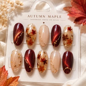 Peut inclure: Un ensemble de dix faux ongles ovales sur une carte de présentation marquée AUTUMN MAPLE HANDMADE PRESS ON NAILS. Les ongles présentent un vernis œil-de-chat bordeaux profond, des bases crème, des feuilles d'or et des motifs d'érables rouges.