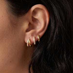 May include: Three gold hoop earrings worn in a single ear. The first earring is a small plain gold hoop, the second is a thin hoop set with small clear stones, and the third is a slightly larger, thicker gold hoop with a polished finish.