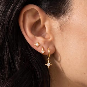 May include: A set of three gold-toned earrings featuring a starburst design with a central clear stone. The top earring is a stud, while the middle and bottom earrings are small hoops, with the bottom hoop featuring a dangling starburst charm.
