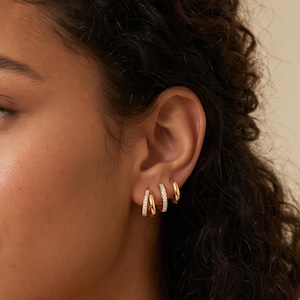 May include: Three small gold hoop earrings worn in a single earlobe. The two front hoops feature a row of small, clear, sparkling stones embedded along the outer edge, while the third hoop at the back is a plain, polished gold band.