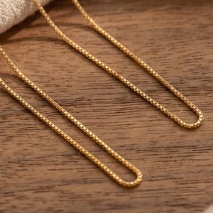 May include: A gold-toned box chain necklace featuring a series of interconnected square links that form a smooth, continuous strand. The metallic chain has a polished finish and is draped in a curved, looping pattern across the frame.