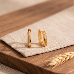 May include: A pair of gold-toned hoop earrings with a rectangular shape and a triple-ribbed texture on the outer surface. The earrings feature a hinged closure mechanism and a polished, reflective finish that catches the light.