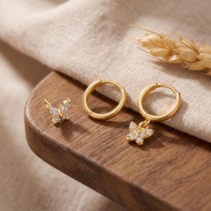 May include: A set of gold-toned jewelry consisting of one butterfly-shaped stud earring with clear stones, one plain gold hoop earring, and one gold hoop earring featuring a dangling butterfly charm encrusted with clear stones.