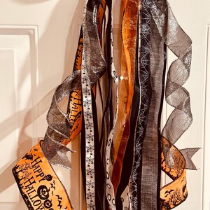 May include: A decorative Halloween bow featuring orange ribbon with black pumpkin and bat prints, black ribbon with white spiderweb patterns, and shimmering silver mesh ribbon. The top bow displays the text Happy Halloween in black lettering.