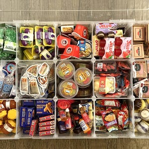 May include: A clear plastic storage container with twelve compartments filled with miniature food items, including Jell-O, Pizza Hut boxes, Auntie Anne's pretzel boxes, small bottles, and various packaged snacks, all organized in a grid layout.