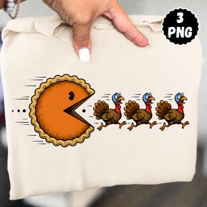 May include: A beige sweatshirt featuring a graphic of an orange pumpkin pie shaped like a video game character chasing three brown turkeys with blue heads and red wattles. The graphic includes motion lines and the text 3 PNG in a black circle.