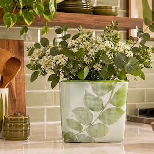 May include: A square glass vase with a light green base color and a dark green leaf pattern created with a dot matrix design. The vase is filled with a lush arrangement of green eucalyptus branches and small white flowers, creating a fresh look.