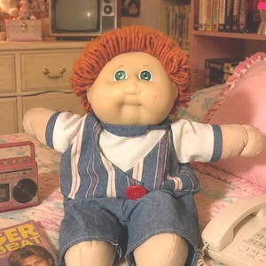 Pode incluir: Uma boneca de corpo macio com cabelo de lã laranja e olhos verdes. A boneca veste uma camiseta branca de manga curta sob um macacão jeans azul com finas listras verticais brancas e vermelhas. Um botão vermelho é visível na frente.