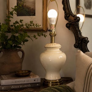 Könnte beinhalten: Eine Tischlampe mit einem glänzenden, cremefarbenen Keramikfuß, einem messingfarbenen Metallhals und einer Harfenhalterung, die eine leuchtende Glühbirne trägt. Die Lampe steht auf einem runden Messingsockel und hat einen abgerundeten, urnenförmigen Körper.
