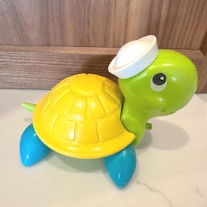 Puede incluir: Un juguete de tortuga de plástico con cabeza y cuello de color verde brillante, un caparazón amarillo con un patrón texturizado y cuatro aletas azules. La tortuga lleva un pequeño sombrero de marinero blanco sobre la cabeza y tiene grandes ojos negros y blancos.
