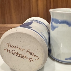 May include: Two ceramic mugs with a white speckled glaze and blue painted accents. The base of the overturned mug features the handwritten text Boulder Pottery M. Cobrado with a small sun symbol etched into the light-colored clay surface.