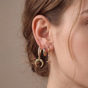 May include: Three gold-tone hoop earrings worn in one ear. The largest hoop features a crescent moon charm with green and clear stones. The middle hoop has a row of multicolored stones. The smallest hoop features a star-shaped drop charm.