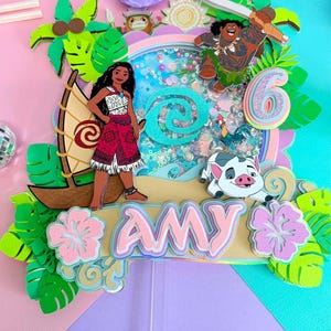 Puede incluir: Un adorno para pastel de papel en capas con los personajes de Moana, Maui y Pua, con un elemento central tipo coctelera lleno de lentejuelas coloridas. El diseño incluye un velero, hojas tropicales, el número seis y una pancarta con el nombre AMY.