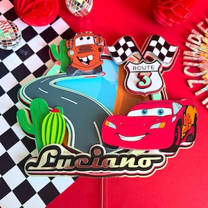 Peut inclure: Une décoration de gâteau superposée représentant une voiture de course rouge avec le numéro 95, une dépanneuse orange, des cactus verts, une route sinueuse et un panneau Route 3 avec des drapeaux à damier. Le nom Luciano est écrit en script.