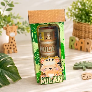 Puede incluir: Una caja de regalo rectangular con tapa marrón texturizada y exterior verde con temática de selva que incluye una ventana transparente. Dentro, un recipiente cilíndrico muestra el nombre MILAN. Un recorte de tigre con el nombre MILAN está fijado al frente.