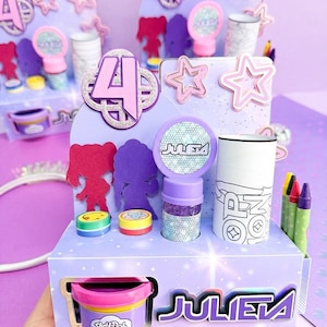 Puede incluir: Una estación de recuerdos de fiesta de cartón de color morado claro con el nombre JULETA en letras negritas. Incluye un recipiente de Play-Doh, un acento circular brillante, un tubo cilíndrico y tres crayones. El expositor está decorado con estrellas recortadas y siluetas de figuras, con el número 4 destacado en la parte superior.