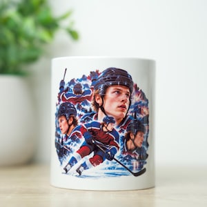 May include: A white ceramic mug featuring a colourful graphic print of a hockey player in various action poses, including skating with a stick and wearing a helmet, set against a backdrop of mountains and a forest landscape in shades of blue and red.