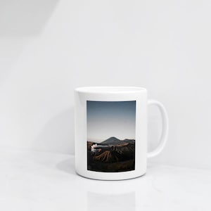 Peut inclure: Un mug en céramique blanche orné d'une impression photographique représentant un paysage volcanique. L'image sur le mug montre une chaîne de montagnes sombre et texturée sous un ciel clair, avec un panache de fumée blanche s'élevant du cratère.