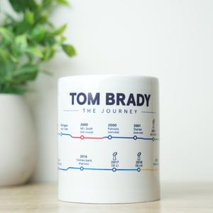 May include: A white ceramic mug featuring a dark blue graphic titled TOM BRADY THE JOURNEY. The design displays a timeline of career milestones with red and blue lines, text labels for years and events, and small trophy icons printed on the surface.