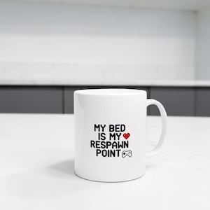 Peut inclure: Un mug en céramique blanche avec le texte noir MY BED IS MY RESPAWN POINT imprimé sur le devant, accompagné d'une icône de cœur rouge et d'une petite icône de manette de jeu noire. Le mug présente une forme cylindrique avec une anse latérale.