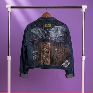 Op de afbeelding: Een donkerblauw spijkerjack met een handgeschilderde Star Wars-scène op de achterkant. Het kunstwerk toont de Millennium Falcon, de Death Star, Han Solo bevroren in carboniet en Chewbacca tegen een sterrenhemel met gele tekst.
