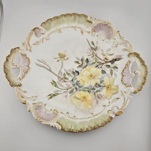 May include: Oval-shaped, white porcelain serving tray with a hand-painted floral design. The tray features yellow roses, green leaves, and brown stems. The edges are trimmed with gold and accented with pale green and lavender details.