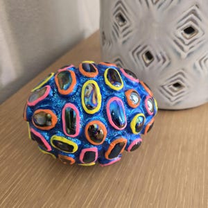 May include: A decorative, spherical object with a blue, glittery surface. It features numerous oval-shaped, iridescent accents outlined in bright colors like pink, orange, and yellow. A white, textured vase is in the background.