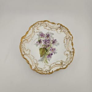 May include: A white porcelain plate with a scalloped, gold-painted edge. The plate features a hand-painted bouquet of purple violets with green leaves. The design is centered on the plate, with gold scrollwork around the edge.
