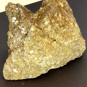 May include: A raw, unpolished mineral specimen featuring a metallic, golden-yellow luster. The rock has a jagged, crystalline surface with numerous small, reflective facets that catch the light, creating a textured, uneven appearance throughout.