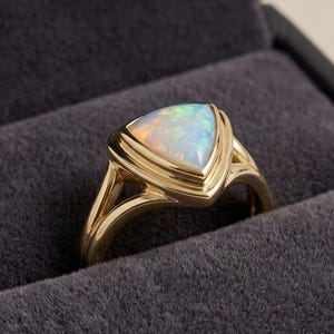 May include: A gold ring featuring a triangular-cut opal gemstone with iridescent flashes of color. The ring has a polished gold band that splits into two sections as it reaches the setting, cradling the stone in a smooth, bezel-style mount.