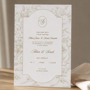 Op de afbeelding: Een witte trouwkaart met een goudkleurige bloemenrand en een centraal ovaal monogram met de letters AJ. De tekst luidt: Together with their families, Adam James & Sarah Elizabeth, Adam & Sarah, Saturday, 15 June 2026.
