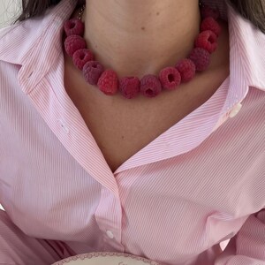 Op de afbeelding: Een ketting met een streng realistische rode frambozenkralen, gedragen bij een lichtroze en wit gestreept overhemd en bijpassende broek. Een wit keramieken bord met een rood bloemmotief bevat een broodje, een klontje boter en wat pasta.