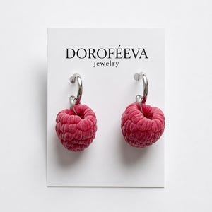 May include: A pair of silver hoop earrings with dangling raspberry-shaped charms on a white display card labeled DOROFÉEVA jewelry. The raspberries are textured and deep pink in color, suspended from polished silver hoops attached to the card.