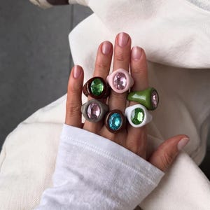 May include: A collection of six chunky rings featuring smooth, solid-coloured bands in shades of brown, pink, olive green, grey, and white. Each ring is set with a large, oval-shaped faceted gemstone in contrasting colours including pink, green, and blue.