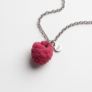 May include: A silver-toned metal chain necklace featuring a realistic, textured raspberry-shaped pendant in a deep pink colour. A small, circular silver charm is attached to the jump ring, clearly engraved with the text DOROFEEVA Jewelry.