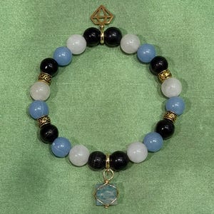 May include: A beaded bracelet featuring alternating black, light blue, and white round beads with gold-toned metal spacers. The bracelet includes a gold-toned geometric diamond-shaped charm at the top and a square light blue stone charm at the bottom.