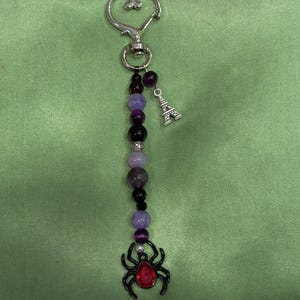 May include: A keychain featuring a silver-toned heart-shaped clasp, a strand of purple and black beads, a small silver Eiffel Tower charm, and a black spider pendant with a red gemstone center. The beads vary in shade and size along the strand.